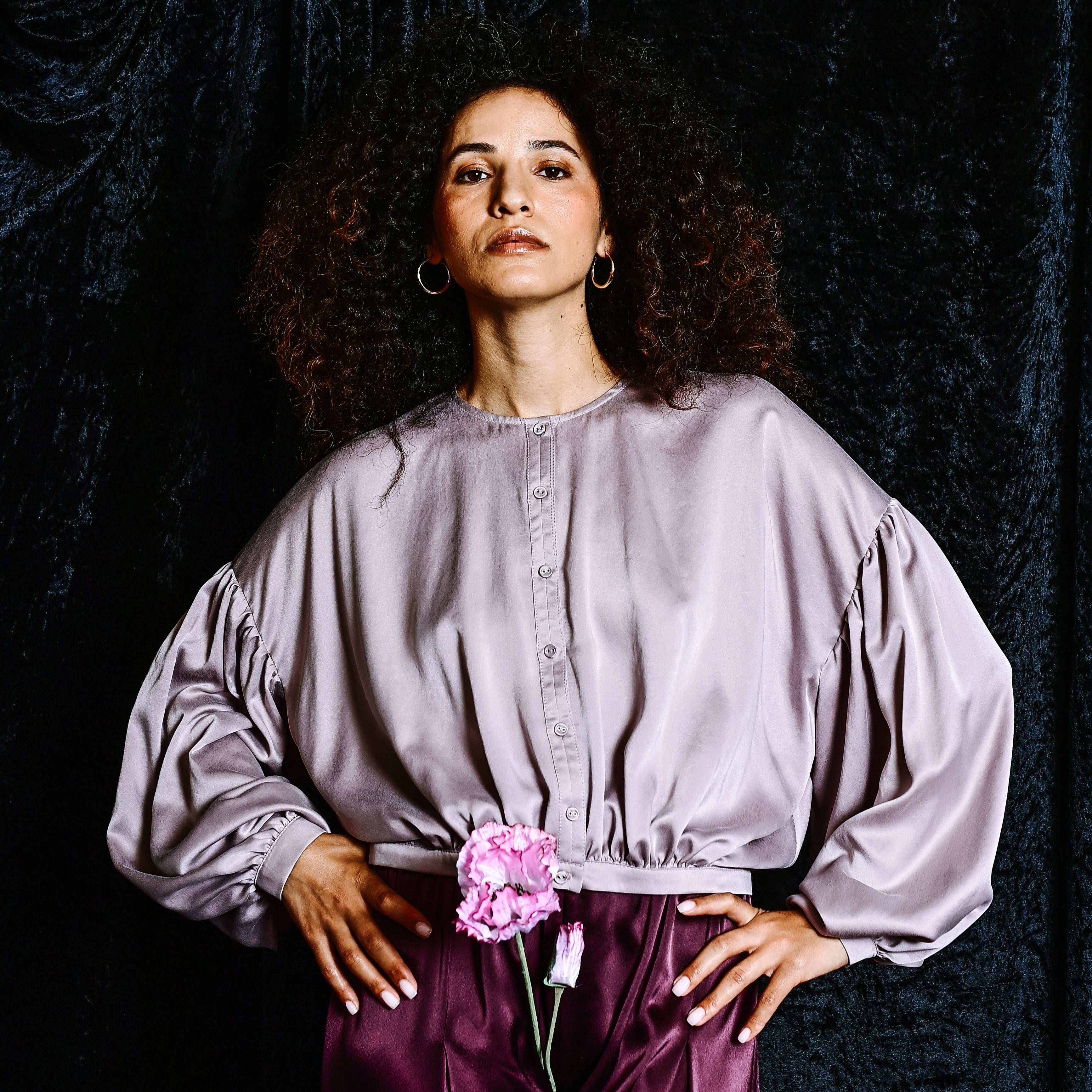 Woman wearing a light purple blouse with puffed sleeves and dark purple pants against a black curtain background.